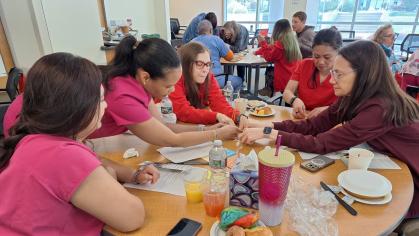 Five women—most wearing bright pink or red scrubs—lean over a round table, carefully assembling a marshmallow‑and‑spaghetti tower during a team‑building exercise. The tabletop is scattered with papers, juice cups, a colorful tumbler, pastries, and other supplies, while additional groups collaborate at nearby tables in the sunlit room behind them.