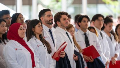 A group of people wearing white coats stand in a row, holding red folders and pamphlets at a ceremony. 