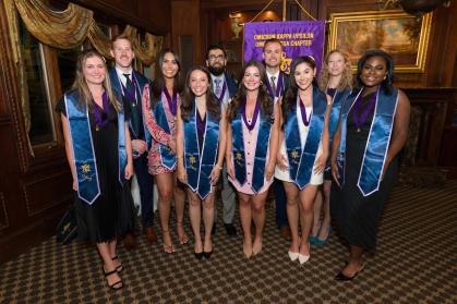 A group of ten graduates stand indoors in formal attire, wearing blue honor stoles with the Omicron Kappa Upsilon (OKU) emblem and purple medallions. They are smiling and posing in front of a purple and gold banner that reads "Omicron Kappa Upsilon, Omega Omega Chapter." The setting appears to be an elegant room with wood paneling, a window with gold curtains, and a framed landscape painting.