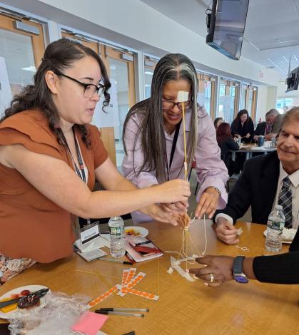 Two women and a man huddle around a round table, steadying a fragile tower made of uncooked spaghetti, tape, and string with a single marshmallow on top during a team‑building exercise; the tabletop is strewn with sticker dots, pens, water bottles, and snack plates, while other groups work in the bright conference room behind them.