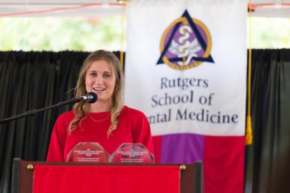 A person in a red shirt stands at a podium, speaking into a microphone. Behind them is a banner displaying the Rutgers School of Dental Medicine logo.