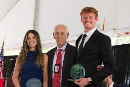 Three individuals stand together in a formal setting. The person on the left, wearing a blue dress, holds an award. The middle person wears a suit with a red tie and an ID badge. The person on the right, in a black suit and white shirt, holds an award labeled.